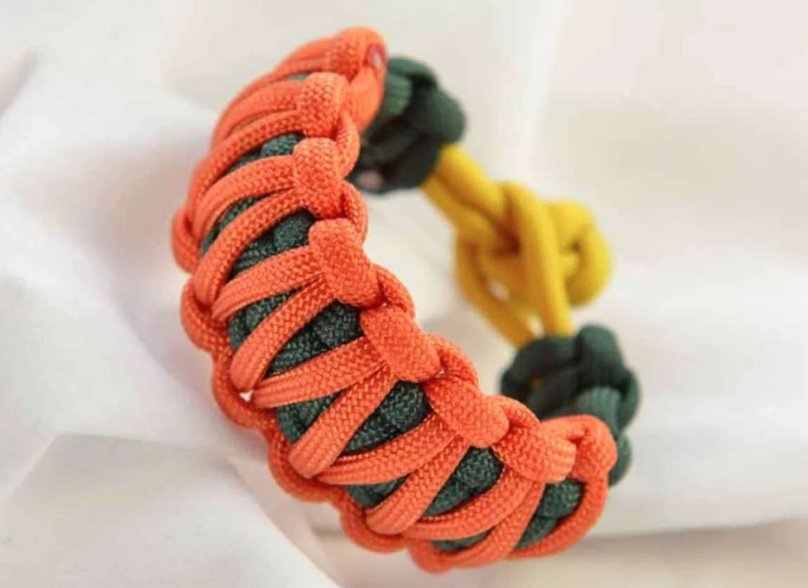 Screenshot 20260708 at 124215 AM Explore Magazine Close-up of a chunky paracord bracelet braided with orange cords over a green core on a white background