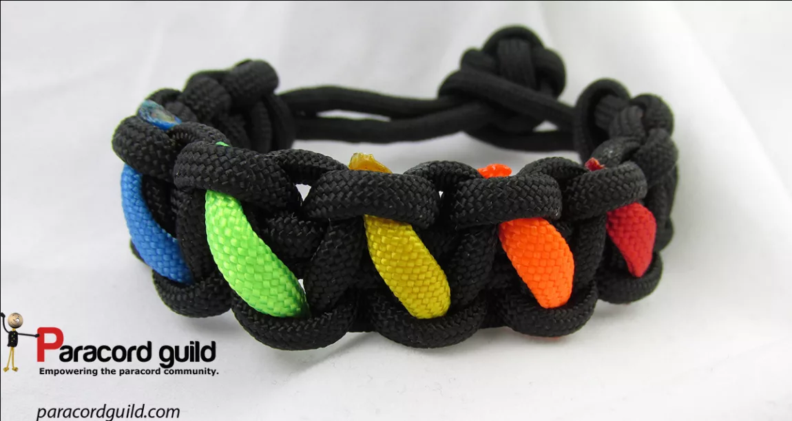 paracordrainbowbracelet Explore Magazine Black paracord bracelet with colorful inner strands (blue, green, yellow, orange, red) on a white background; Paracord Guild product image with logo in corner.