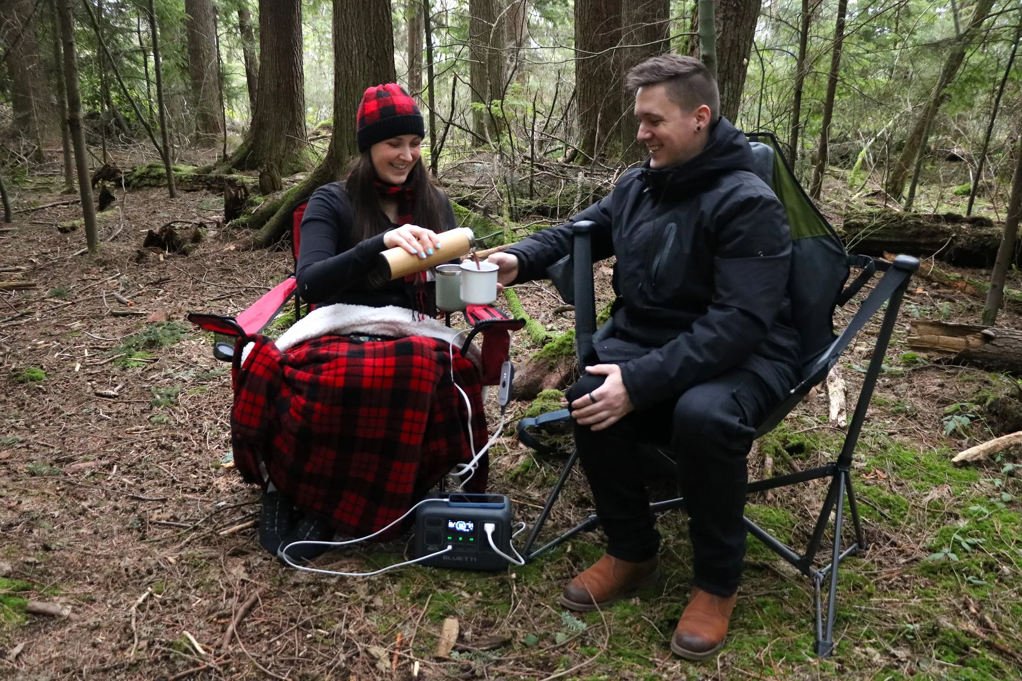 Two people outdoors enjoying the forest while staying warm with a heated blanket and hot chocolate. Yum!