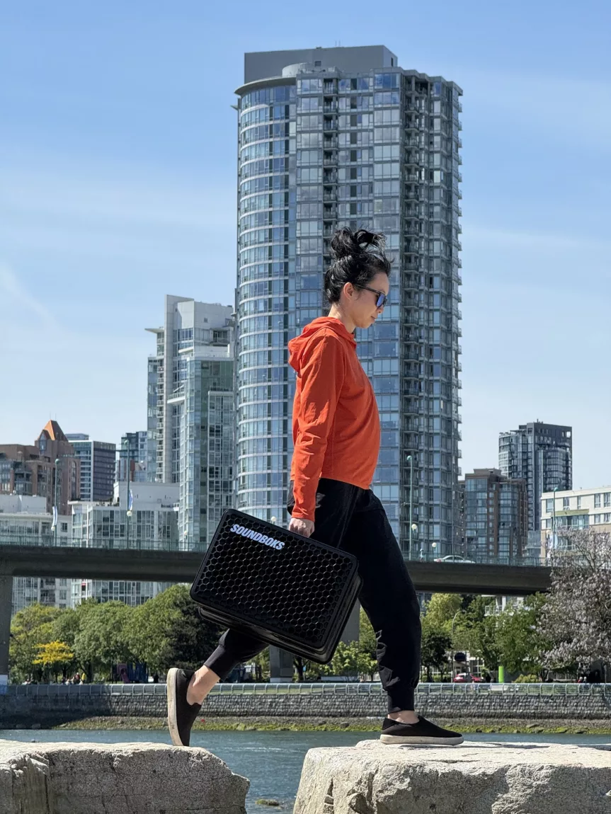 Woman carrying speaker outside in a park on grass in front of a large building in the city.