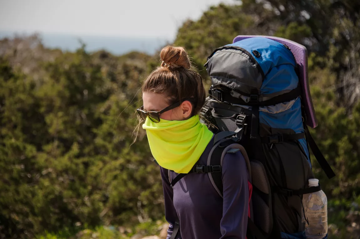 Explore Magazine Girl standing with a hiking backpack and special mask from dust Explore Magazine Girl standing with a hiking backpack and special mask from dust on the background of green trees and clear sky