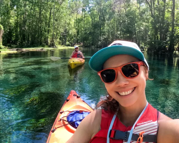 Smiling woman in a red life vest and teal cap in the foreground, kayaking on a clear green river with another person in a yellow kayak in the background among trees.