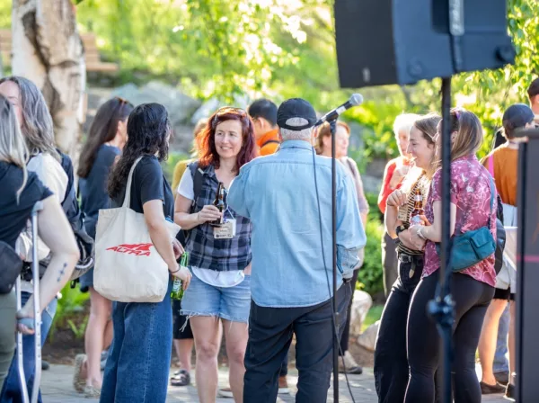 Group of friends at an outdoor gathering, chatting and holding drinks near a microphone and speaker.