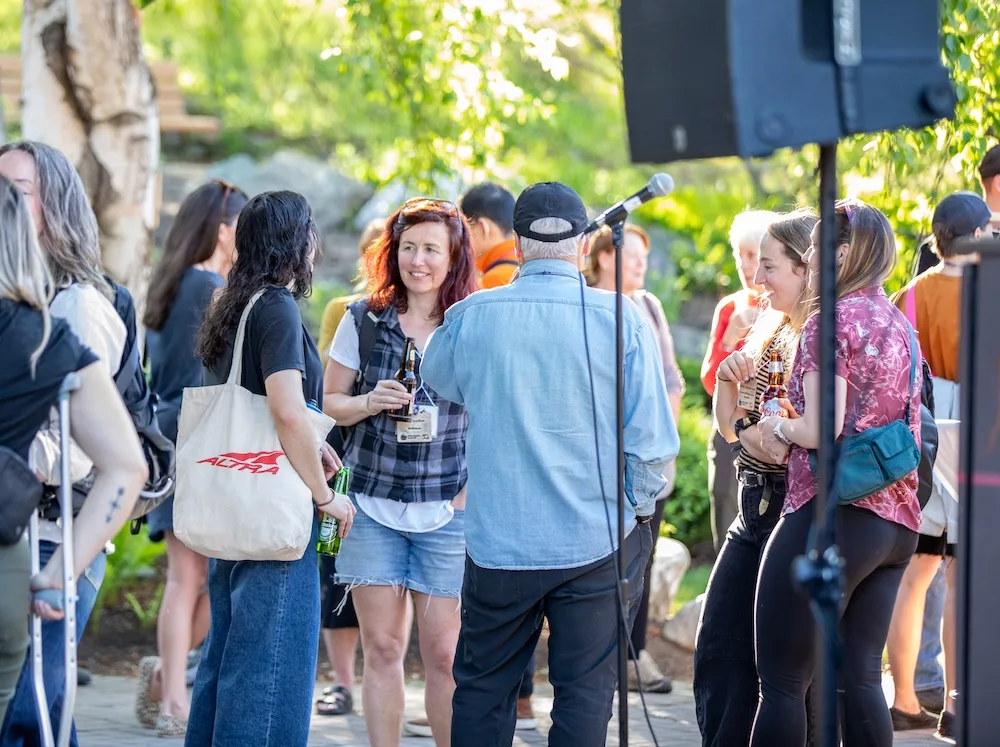 Group of friends at an outdoor gathering, chatting and holding drinks near a microphone and speaker.