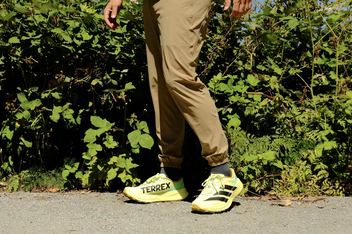Person's legs in beige pants and bright yellow Terrex sneakers stepping on a gravel path with green foliage in the background.