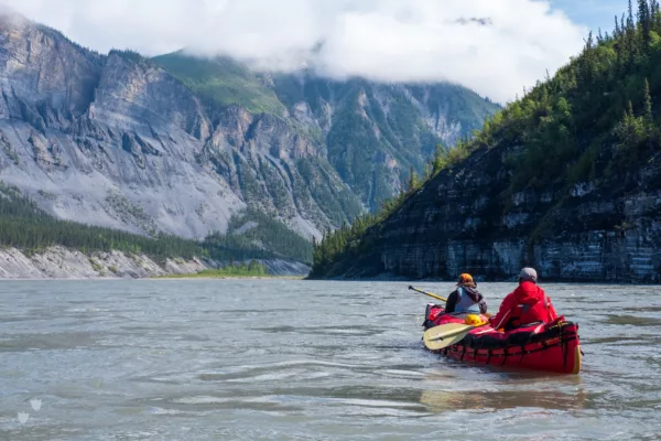 Two people in a red inflatable raft paddling along a wide river with steep rocky cliffs and forested mountains in the distance.