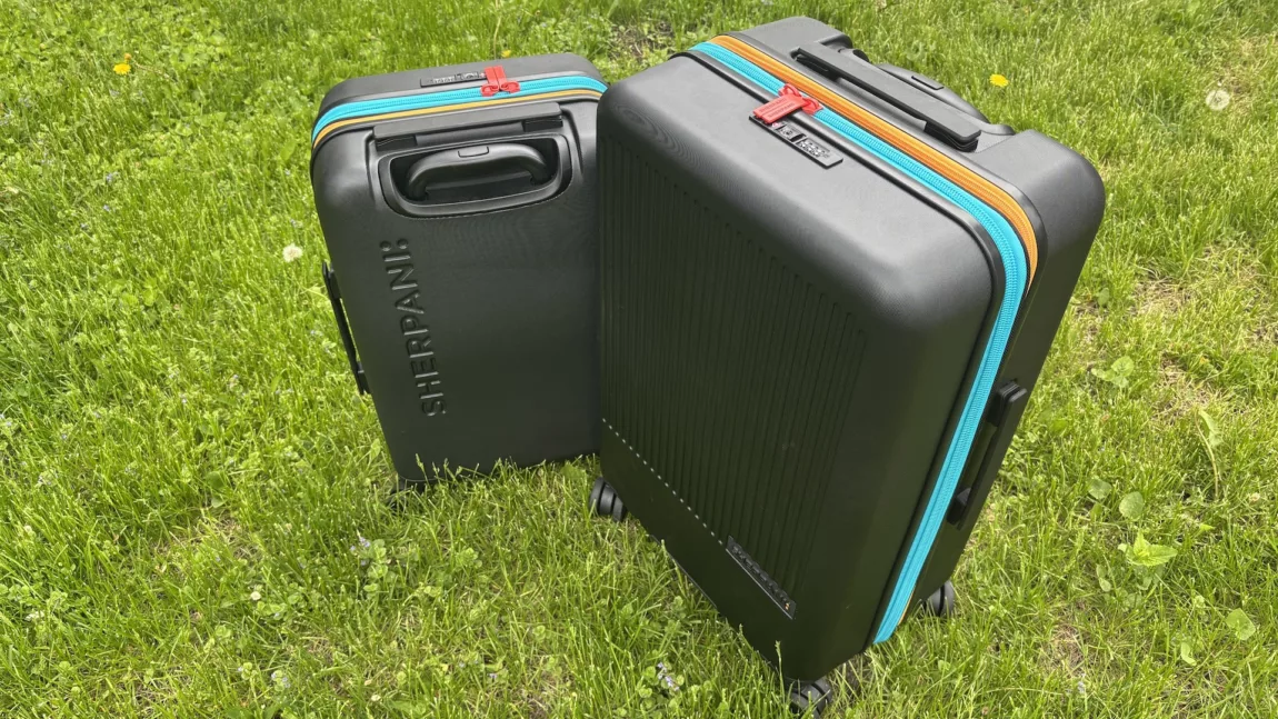 Two black hard-shell suitcases with bright turquoise and orange zippers standing upright on a grassy field on a sunny day.