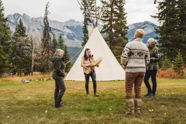 Group of adults standing in a grassy campsite near a white tipi, a woman holding a drum, mountains in the background with trees around.