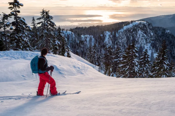 Skier wearing red pants and a blue backpack stands on a snowy slope, gazing at snow-covered mountains and a sunset over a distant bay.