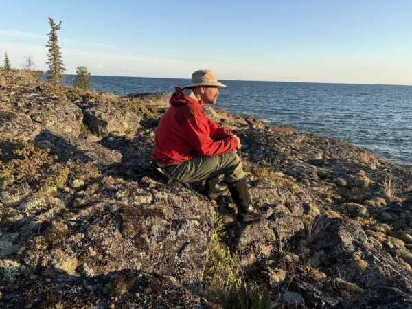Man in a red jacket crouches on rocky shore overlooking the sea, wearing a beige hat and hiking boots