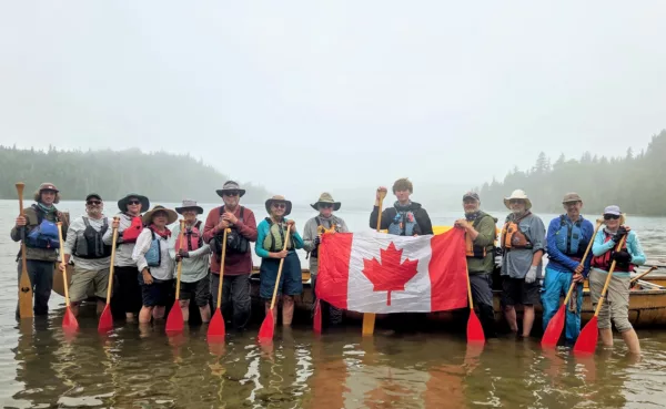 Group of people in life jackets standing in shallow water with paddles, holding a Canadian flag across a canoe/boat backdrop.