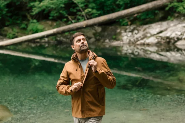 Man in a brown jacket standing by a clear green lake, looking up toward the trees in a forested area.