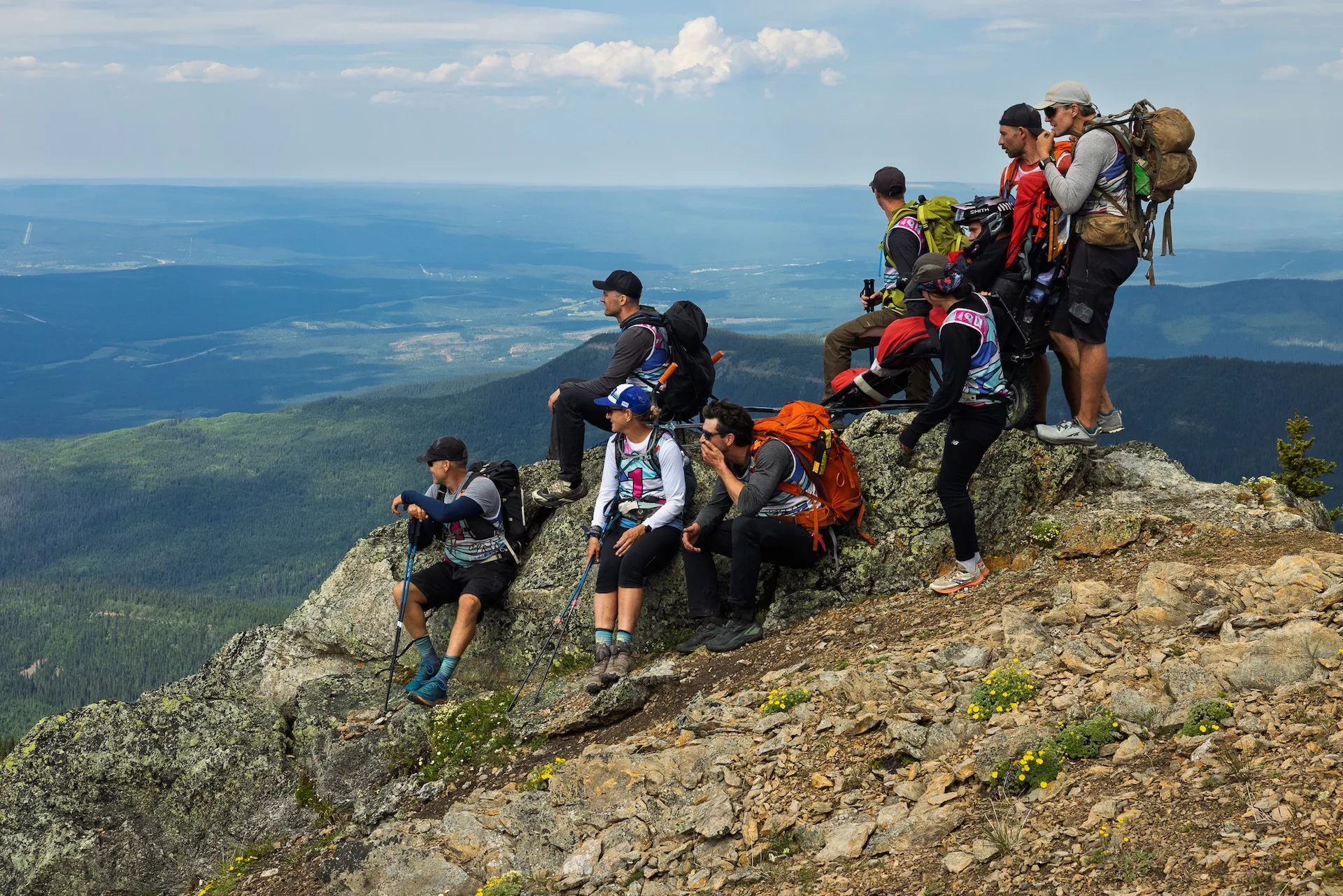 Group of hikers with backpacks and trekking poles standing on rocky cliff, overlooking a valley and forested mountains.