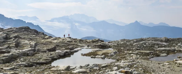 Two hikers in red jackets stand on a rocky mountain ridge with snow-capped peaks in the distance, under a pale blue sky.