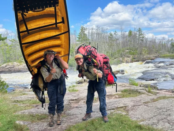 Two hikers with large backpacks pose on a rocky riverbank, a yellow canoe is held overhead between them beneath a blue sky.]}}``` Wait ensure proper JSON. Let's craft properly. } with array 3 items. Let's finalize. )''»} Actually I'll provide three alt texts. Ensure quotes. Let's output. } 0? We'll produce: {