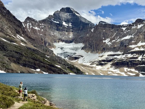 Lake McArthur Lake OHara Explore Magazine Two hikers on a rocky lakeshore overlook a turquoise alpine lake with snow-dusted, jagged mountains behind them.
