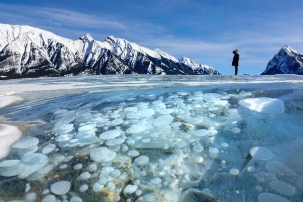 CLOSE UP Woman on trip across Canada marvels at the icy lake under the Rockies Explore Magazine Person standing on a flat icy surface with snow-covered mountains behind and a frozen lake filled with circular blue ice bubbles in the foreground.