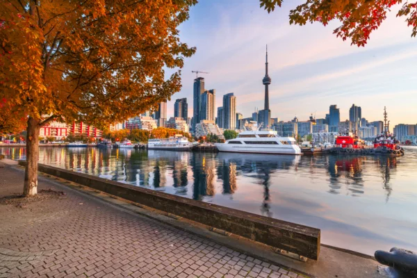 Toronto Ontario Canada Downtown Cityscape at Twilight Explore Magazine Autumn scene at the Toronto waterfront: orange-leaved trees along a paved promenade, with boats and a city skyline including the CN Tower reflected in calm water.